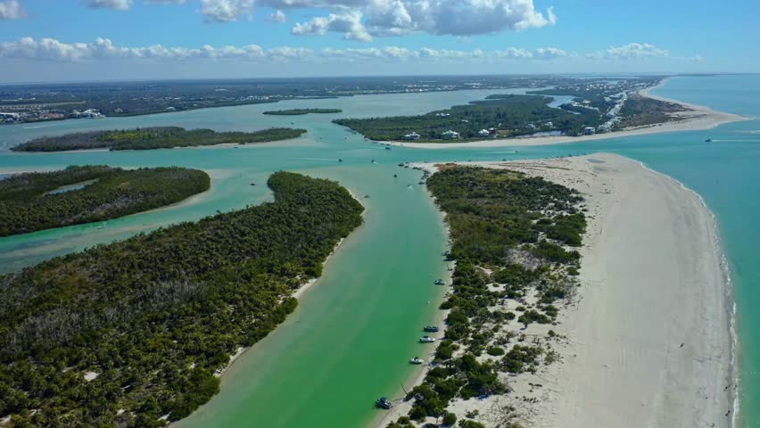A broad coastal inlet running between barrier islands in Florida with anchored boats along the riverbank, showcasing a mix of dense vegetation, sandy shores, and calm turquoise water under a cloudy