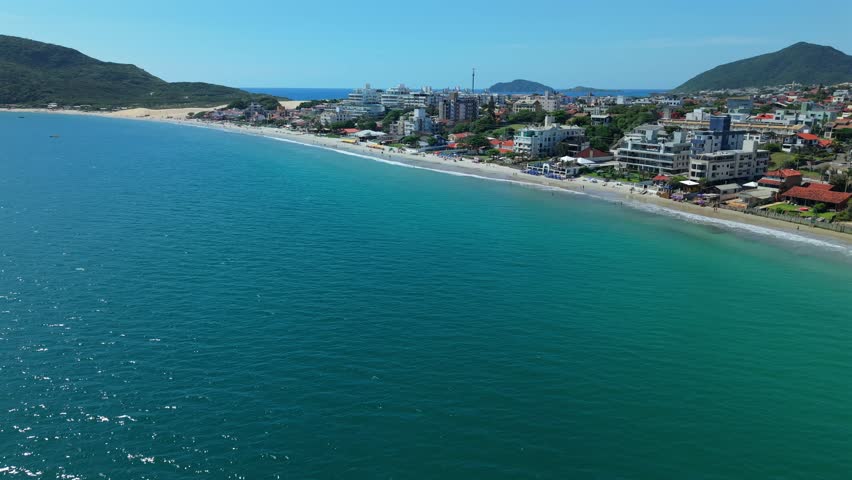 Drone flying forward from turquoise ocean toward crowded beach coastline featuring luxury hotels green mountains and sunny blue sky in Santa Catarina Brazil