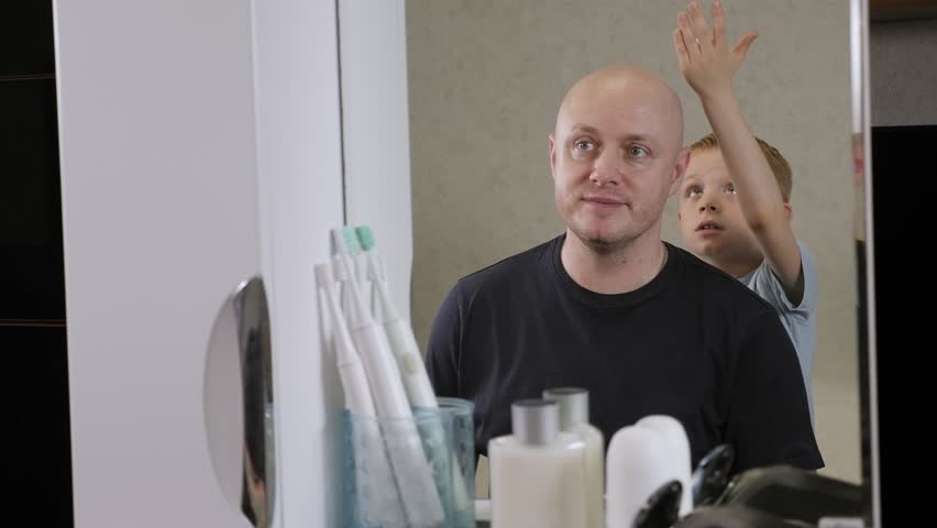 A cheerful little boy applies shaving cream on his father's head in the bathroom in front of the mirror.