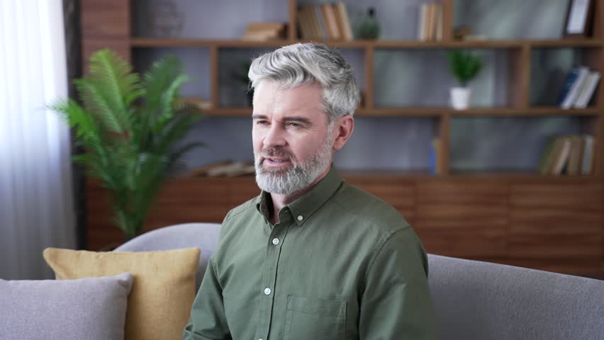 Close-up of smiling mature man relaxing with eyes closed and hands behind head at home. Happy male resting on sofa in cozy living room, enjoying peace, comfort, and pleasant relaxation after work