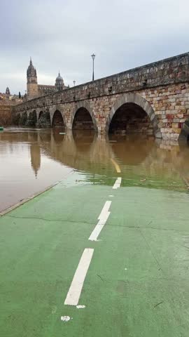 Overflowing river flooding park path and trees near old stone bridge