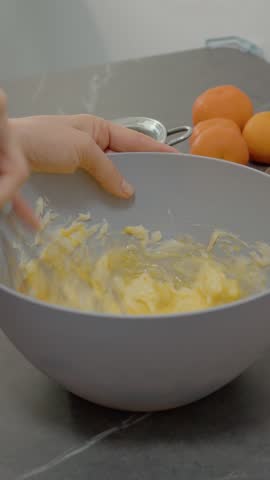 Vertical Screen: A woman expertly combines egg and butter in a bowl, preparing for a tasty baking session in her warm kitchen filled with fresh oranges.