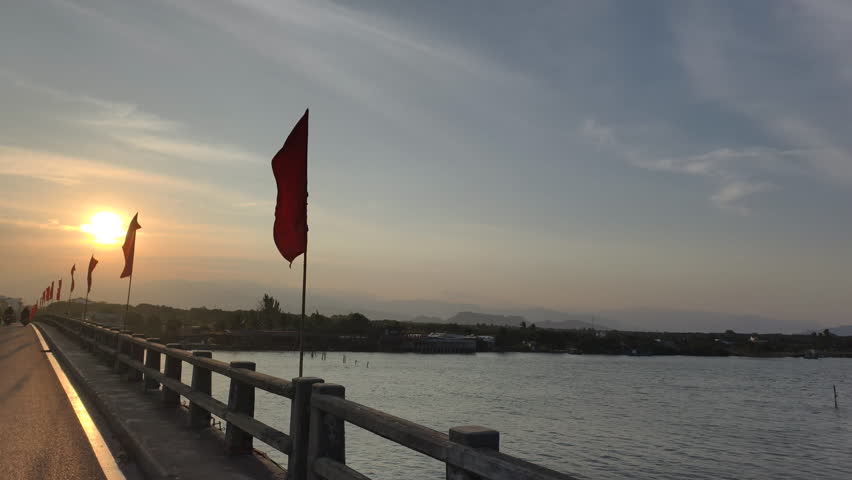 Patriotic Vietnam Bridge features long concrete roadway decorated with fluttering red flags during sunset. Scenic river crossing highlights national pride on Vietnam Bridge under golden evening sky.