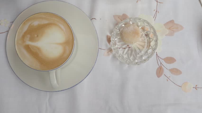 Top-down shot of coffee cup with heart latte art on white tablecloth with small glass bowl holding cookie
