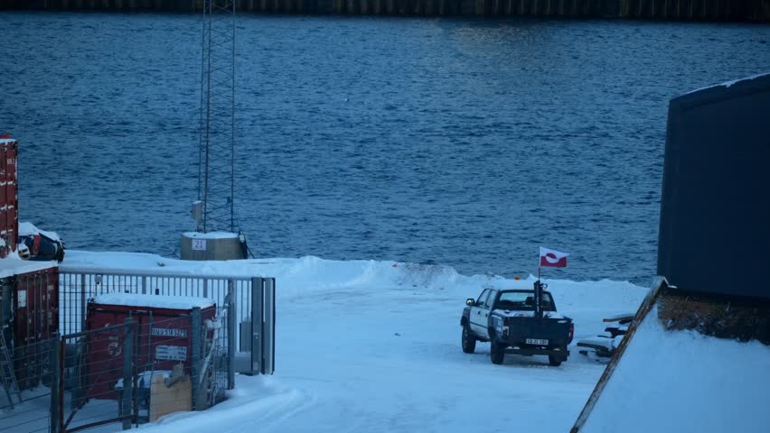 A black pickup truck, the Greenlandic flag stands on a snowy pier. The deep blue waters of a fjord or harbor ripple in the background of this cold coastal scene.