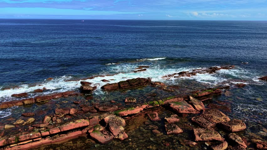 Beautiful seascape waves meeting layered red rocks on North Sea coast in Scotland. Swells washing over weathered formations in shallow water. Foam trailing across sandstone ledges under blue sky