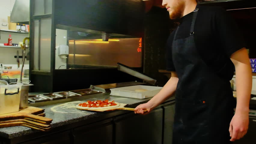 Man chef carefully places prepared pizza into oven to begin baking process. Cook places pizza inside wood-burning oven. Slow motion