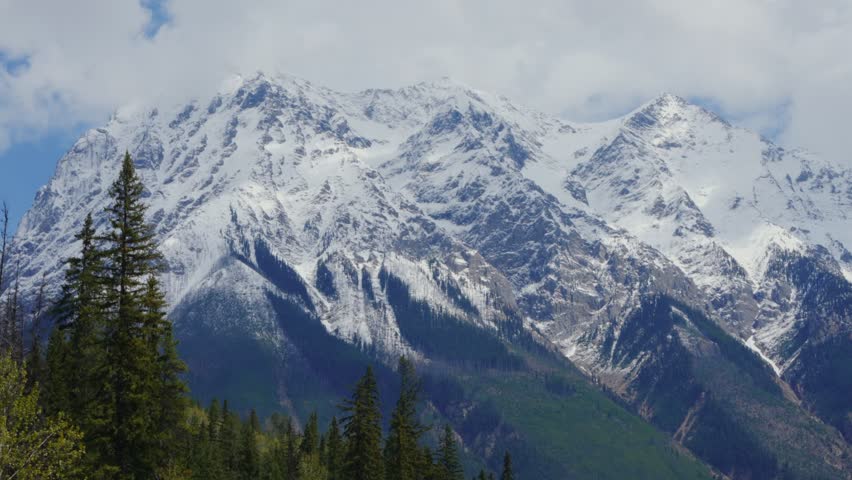 Snow capped peaks of Yoho National Park standing tall under a cloudy sky with green forest at the base