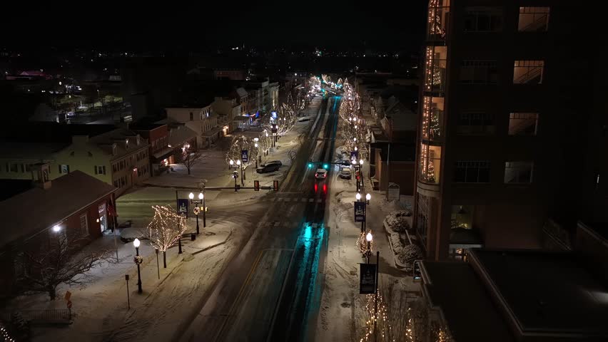 Aerial night view of a snowy American street with Christmas lights, illuminated trees, quiet traffic, and warm residential windows, showing winter atmosphere in small town of USA. Flyover.