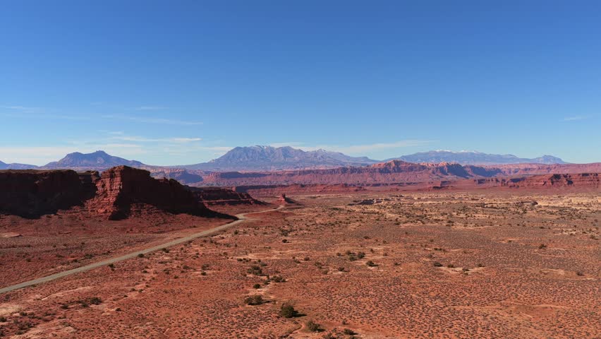 A wide drone shot captures a lonely paved road winding through alien red rock formations and arid desert basins. This scifi Utah landscape features mystical mesas and a vast, hazy horizon.