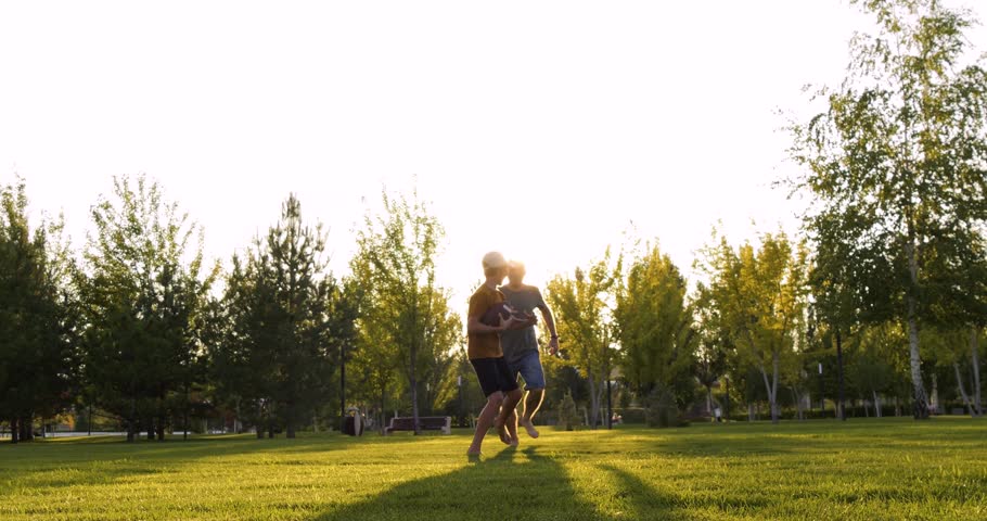 Active father chases running son with rugby ball. Sporty father races with boy in sunny park. Happy father plays American football game. Outdoor family fitness and cardio sport concept. Dad and kid