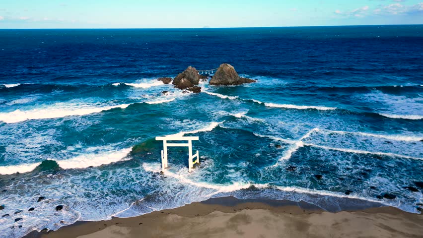 Aerial view of traditional Japanese torii gate by the sea with rocky formations in the background. Ocean waves create dynamic coastal scene with spiritual landmark.