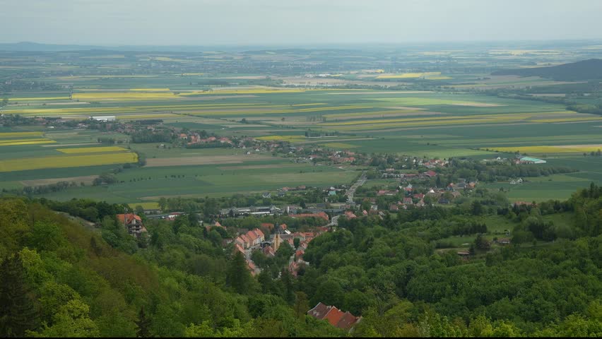 A panoramic view of farmland and a small village spread across rolling hills. The organized fields and open sky give a peaceful rural charm.