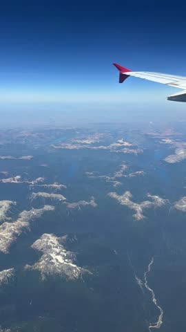 Vertical aerial view of the Argentine Lake District and Patagonian Andes, seen from a commercial airplane window during flight over southern Argentina. Mountain ridges, winding lakes, and dense forest stretch across the landscape beneath clear blue skies, capturing the vast scale and remoteness of Patagonia from above.