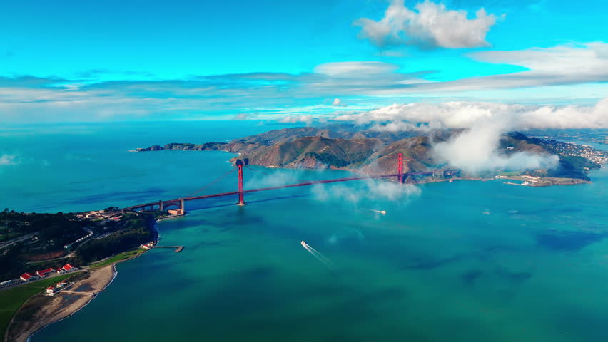 Wide panoramic aerial view of the bay with the Golden Gate Bridge and moving cars. Turquoise ocean waters in sunny weather with small clouds. View from the city of San Francisco.