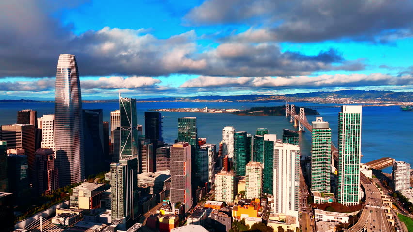 Aerial perspective of the downtown San Francisco skyline, featuring modern skyscrapers and the Bay Bridge in the background under a partly cloudy sky. The ocean is filled with light ripples.