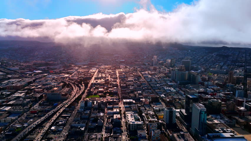 High-angle aerial view of San Francisco city grid with sunset light illuminating the streets. A thick layer of clouds is hovering over the hills in the background. Dense traffic on the freeway.