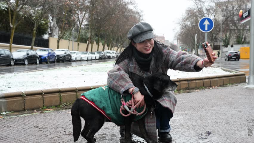 Madrid snowstorm selfie: Smiling woman takes smartphone photo with black senior dog wearing green coat. Happy pet owner enjoying winter weather in the Spanish capital.
