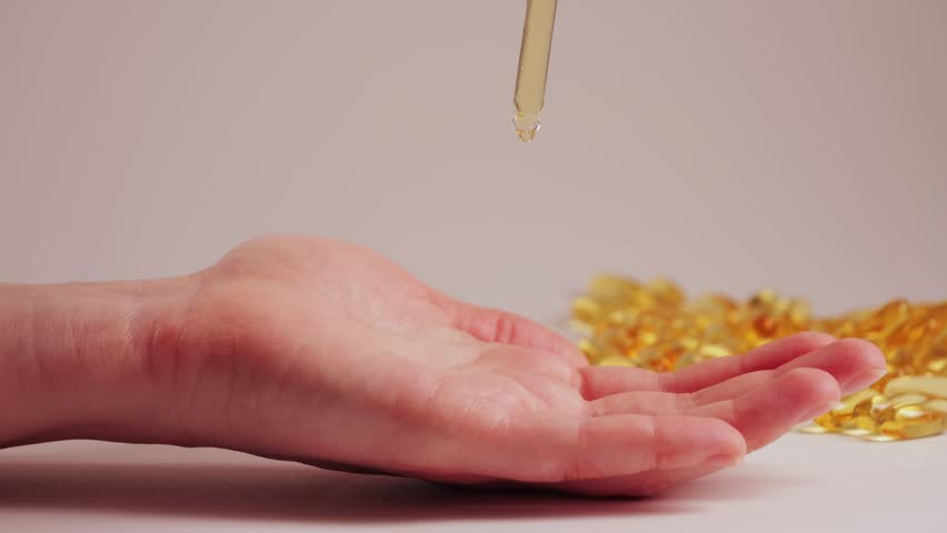 A pipette with golden oil drips onto an open palm. In the background, capsules, vitamins, fish oil, and omega 3 are shown. Health care concept: dietary supplements, natural oils, and cosmetology. High