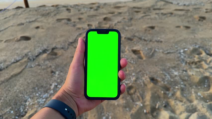 A hand holding a smartphone with a green screen against a sand beach background at sunset during vacation. High quality 4k footage