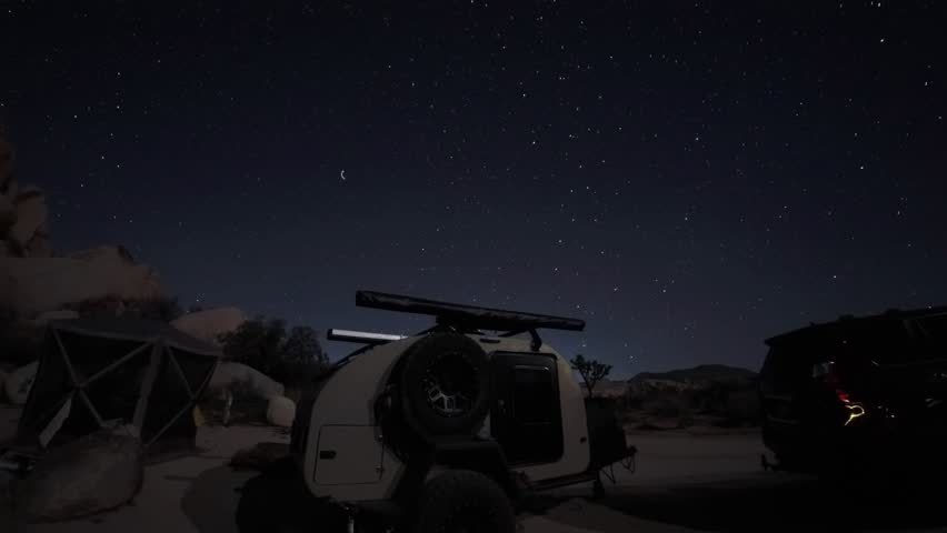 Teardrop camper sits at a campsite while star trails move across the night sky above Joshua Tree National Park in California.