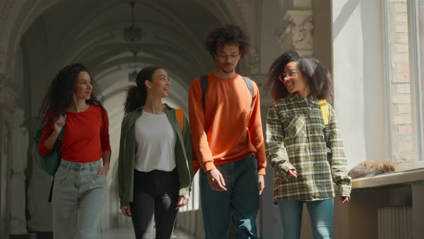Four multiracial students walking together talking in university corridor hall multiethnic friends walk African American man male guy with three women girls females classmates go to lecture education
