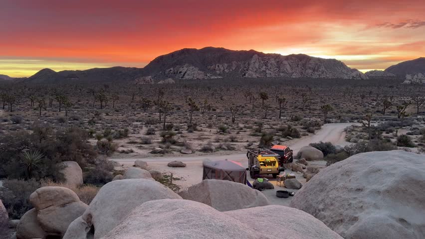 Campers enjoy sunrise in Joshua Tree National Park with a teardrop trailer. The sky is orange red over the desert landscape.