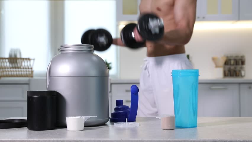 Man exercising with dumbbells, protein powder, shaker and jars in kitchen, closeup