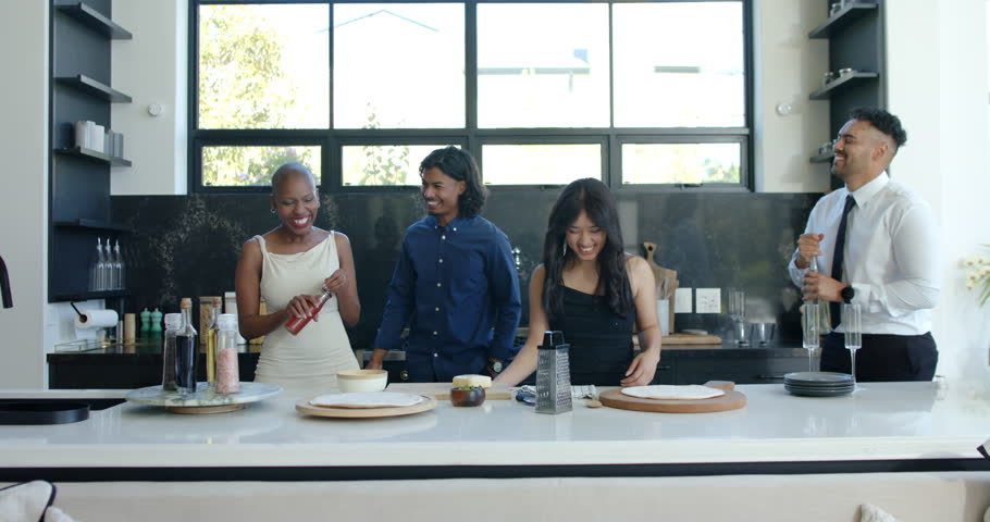 Diverse friends at kitchen island plating while rightmost opening clear bottle, spilling, laughing. Group, gathering, modern, bright, casual, hospitality, culinary