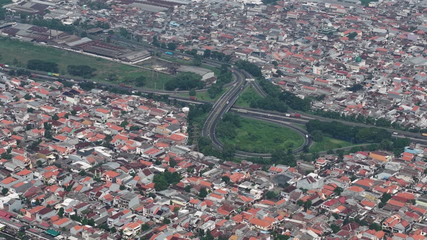 Aerial shot of Surabaya showing dense urban housing, red-tiled rooftops, and a large circular highway interchange surrounded by green space. Captures city life, transportation, and suburban patterns.