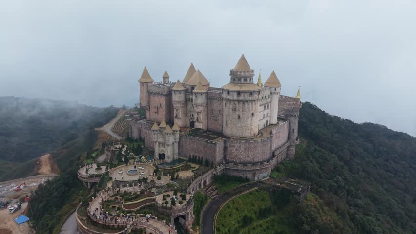 Aerial Misty Castle On Mountain Ba Na Hills Vietnam, Drone Survey Capturing Medieval Facade, Winding Ornamental Gardens, Stone Ramparts, Dense Cloud Cover, Moody Cinematic Atmosphere,