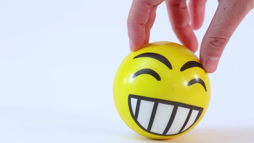 A hand spins a yellow laughing emoji stress ball on a plain white surface