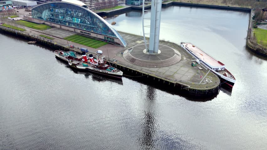 Aerial drone footage descending toward the historic paddle steamer Waverley moored beside the Riverside Museum on the River Clyde in Glasgow, Scotland, showing the waterfront and modern museum architecture from above.
