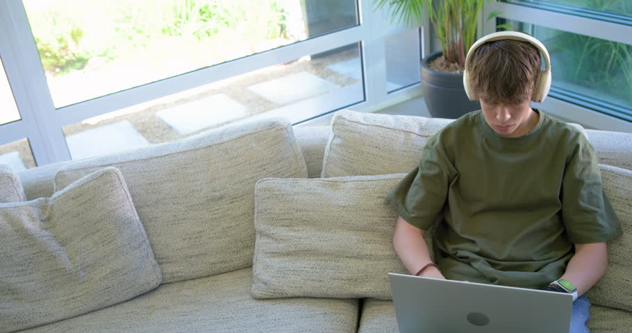 Adolescent male sitting on sofa near window looking down typing on laptop reacting to content. Headphones, computer, smartwatch, sunlit, home, youth, plant