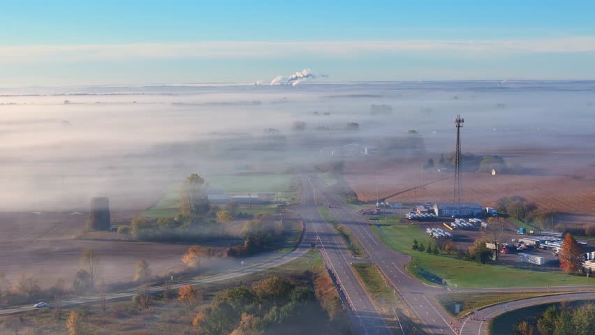 Lake Odessa, Michigan, USA in morning light with fog covering land and roads in aerial view, interstate highway intersection with vehicles and a communication tower visible