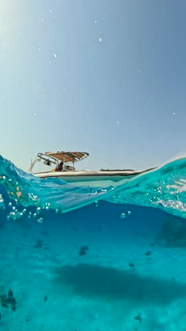 Half underwater vertical view of motorboat anchored in crystal clear sea with waves and bright sunlight