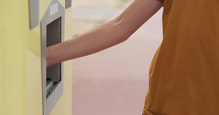 Teenager fills cup with clean water at dispenser. Child drinks fresh water near yellow machine. Boy needs cool water for hydration. Outdoor station pours liquid. Hand holds white container in city.