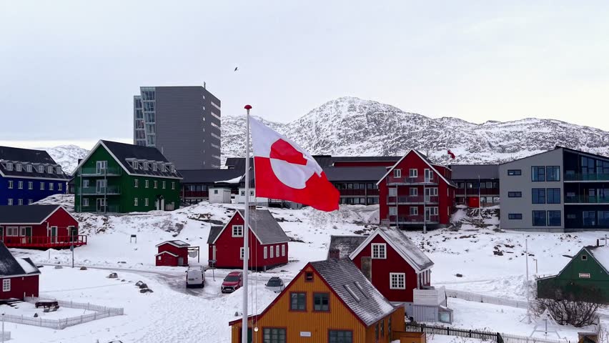 The red and white Greenlandic flag (Erfalasorput) billows in the wind with colorful Nordic houses and snow-capped mountains in the background during winter.