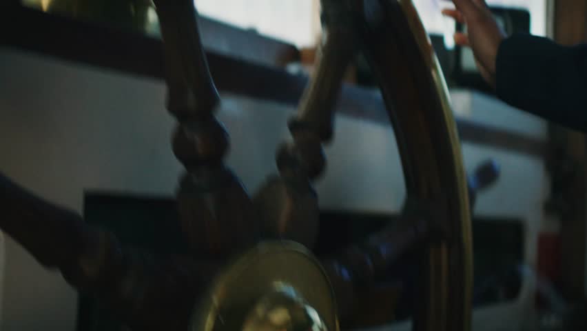 Handheld close-up of boat steering wheel with hand gripping wooden helm, natural light through window, maritime navigation and control concept.