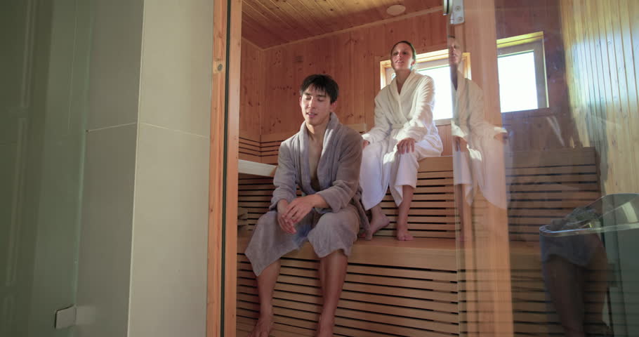 Couple in bathrobes sitting on wooden sauna benches, breathing deeply and reacting to sauna heat. Wellness, spa, steam, warmth, relaxation, rustic, window