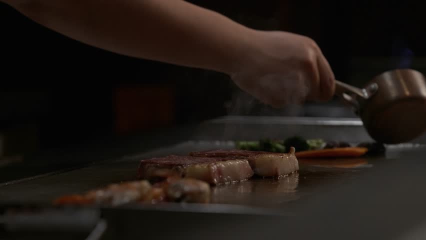 Chef cooking on hot griddle with dramatic slow motion flare-up, hand holding small saucepan over sizzling meat in restaurant kitchen, warm low light, action close-up.