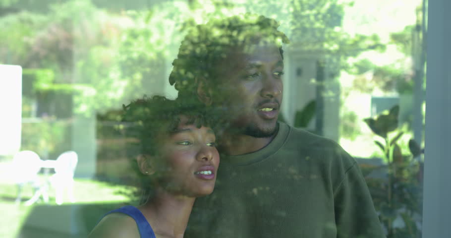 African American couple standing at window watching sunny yard, woman humming and leaning into man. Duo, romance, affection, intimate, sunlight, reflections, glass