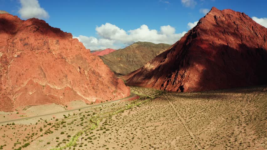 Aerial ascend of towering red cliffs and open desert near Ruta de los Seis Miles divided by road, cloud shadows across cliffs, Catamarca Argentina
