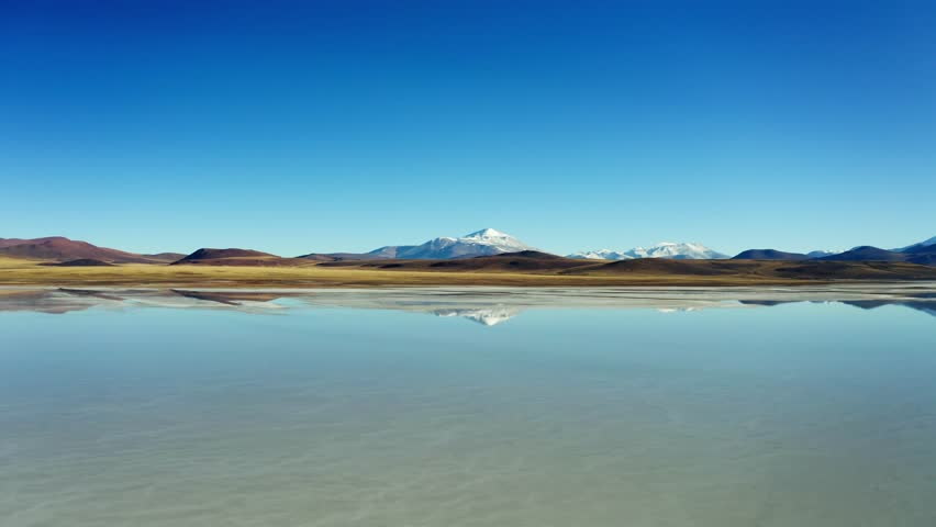 Wide aerial dolly of glassy lake surface in high desert environment reflecting snow mountains under blue sky, Laguna Brava, La Rioja Argentina