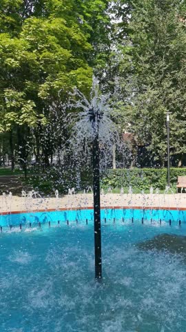 A fountain in a city park. Streams of water cascade against a backdrop of green trees on a summer day. Vertical video.
