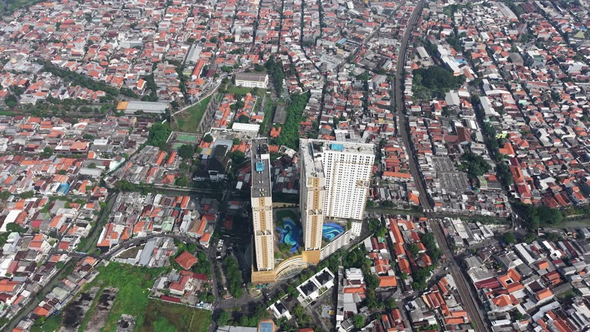 An aerial perspective over a cluster of high-rise residential towers in Surabaya, bordered by a dense mosaic of low-rise homes. The scene captures urban density, modern housing, and city life.