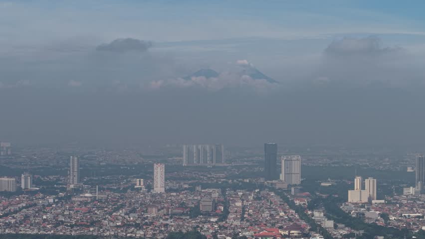 A expansive cityscape of Surabaya features a densely packed downtown with high-rise buildings, set beneath a hazy, dusk sky and distant mountain backdrop for a calm, metropolitan mood.