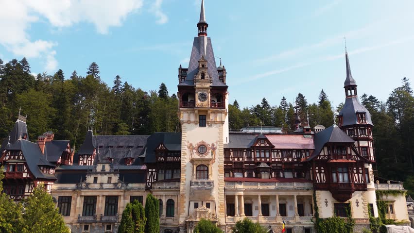 Drone pan revealing Peleș Castle with its central tower framed by pine forest.