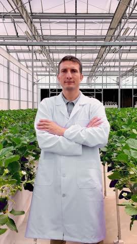 A researcher in a white lab coat stands among rows of leafy plants inside a modern greenhouse, suggesting controlled-environment agriculture and crop research