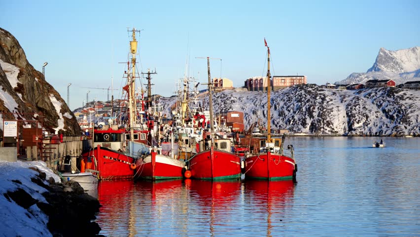 A line of iconic red wooden fishing vessels moored at a pier in Nuuk, Greenland. The background features colorful arctic houses and snow-covered mountains under a clear sky.